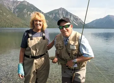 Stephanie and Mike Trost standing in a lake in waders, Mike holding a fly rod, mountains behind them